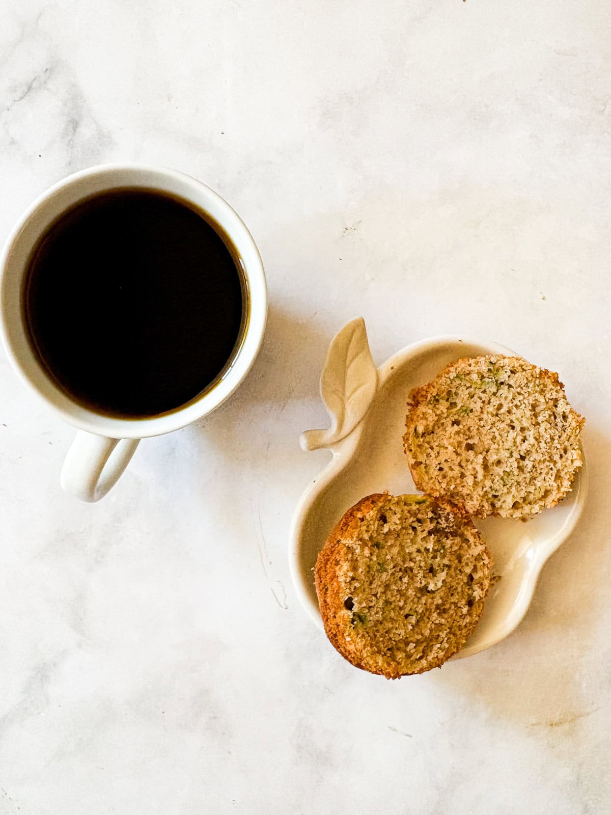 Tuck shredded squash into these gluten free zucchini chocolate chip muffins for a delectable morning treat. A gluten free zucchini chocolate chip muffin cut in a half on a plate and a cup of coffee.