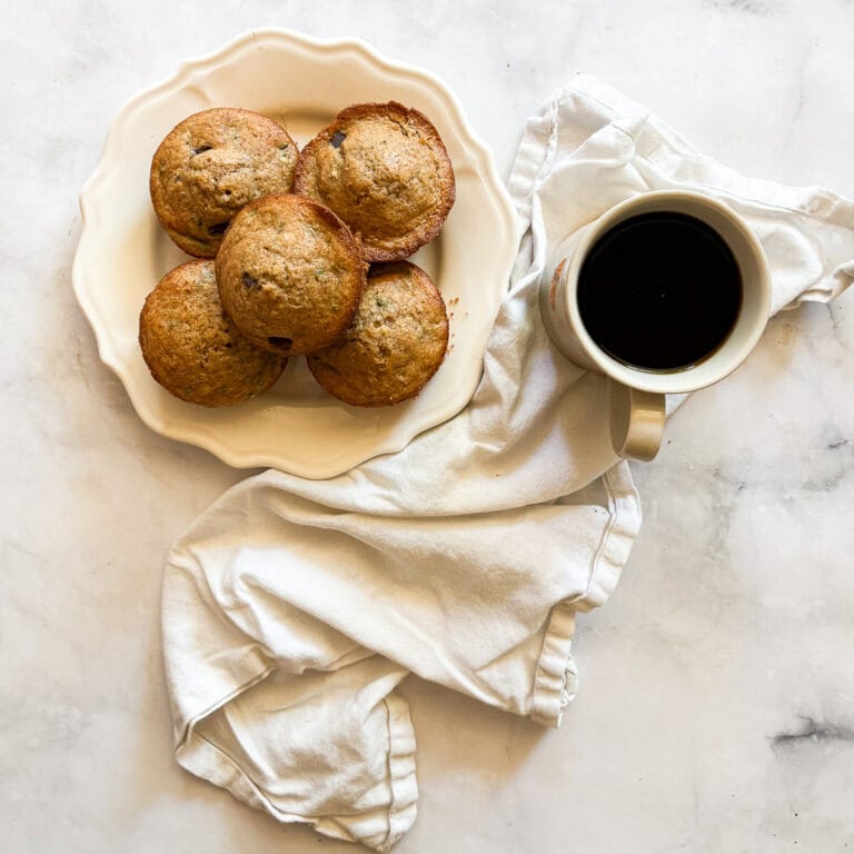 A cup of coffee on a napkin next to a plate of gluten free zucchini muffins.