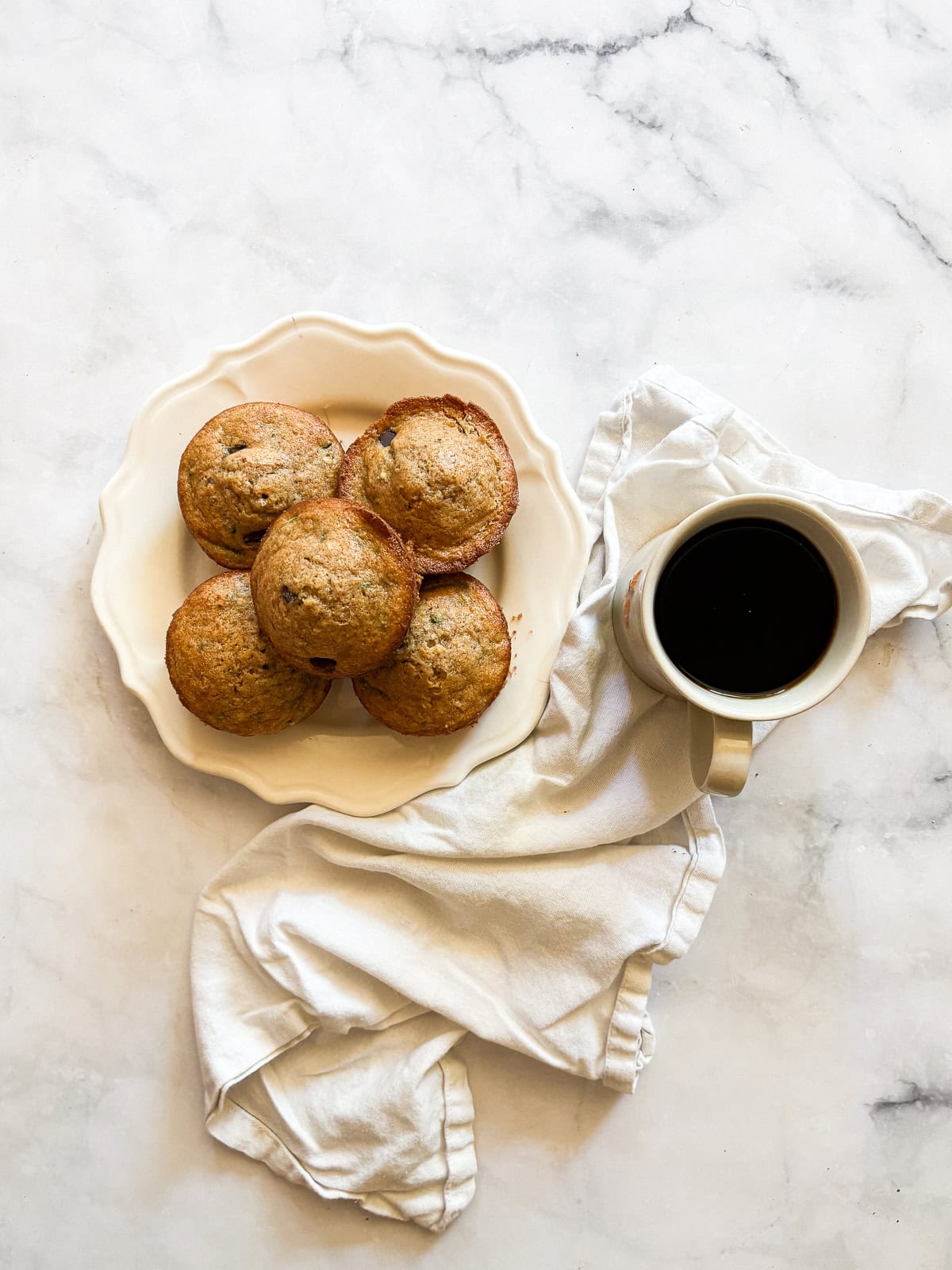 A plate of gluten free zucchini muffins is served next to a cup of coffee and a napkin.
