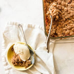 A bowl of gluten free apple crumble topped with ice cream next to a pan of crumble with a serving spoon.