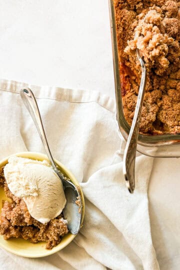 A bowl of gluten free apple crumble topped with ice cream next to a pan of crumble with a serving spoon.