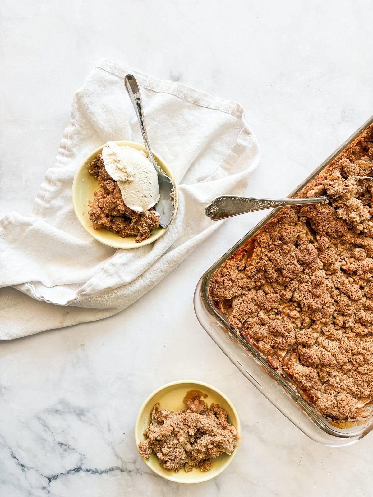 A crisp, buttery brown sugar streusel tops tender apples in this homey gluten free apple crumble. Perfect with vanilla ice cream! Two bowls of gluten free apple crumble, one topped with ice cream, next to a crumble.