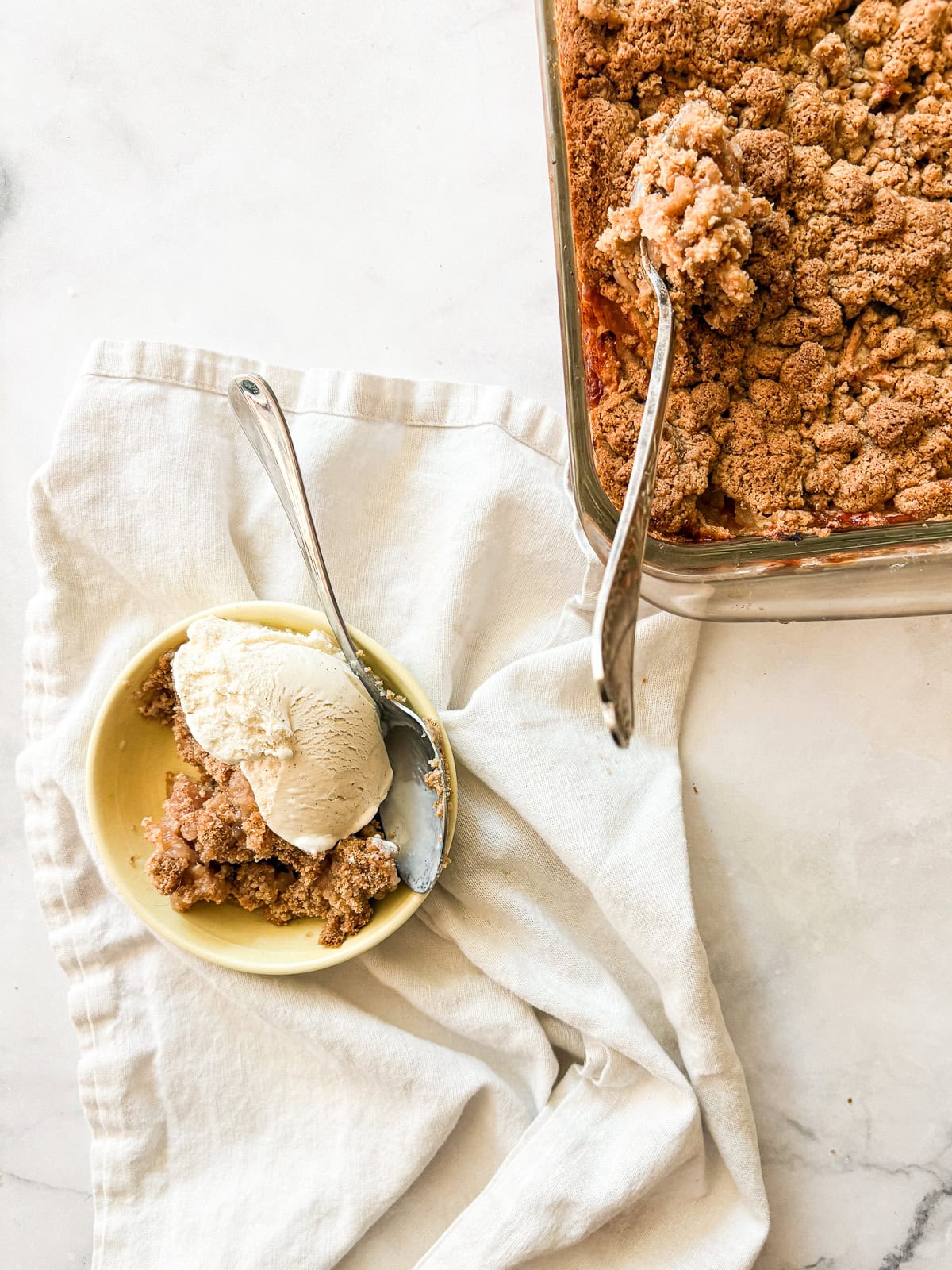 A crisp, buttery brown sugar streusel tops tender apples in this homey gluten free apple crumble. Perfect with vanilla ice cream! A bowl of gluten free apple crumble topped with ice cream next to a pan of crumble with a serving spoon.