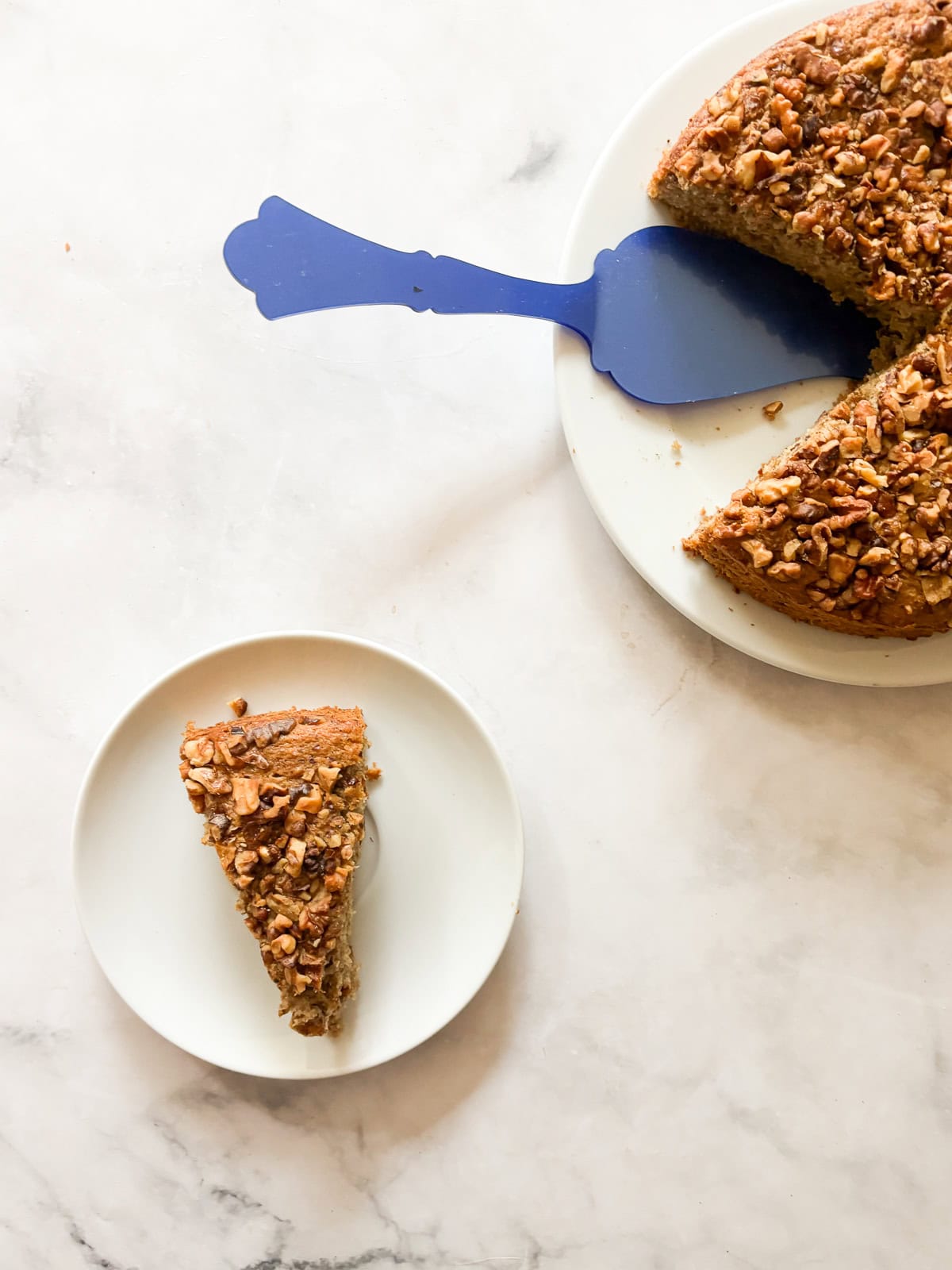 A blue server rests on a plate of gluten free banana walnut cake, with a slice on a plate next to it.