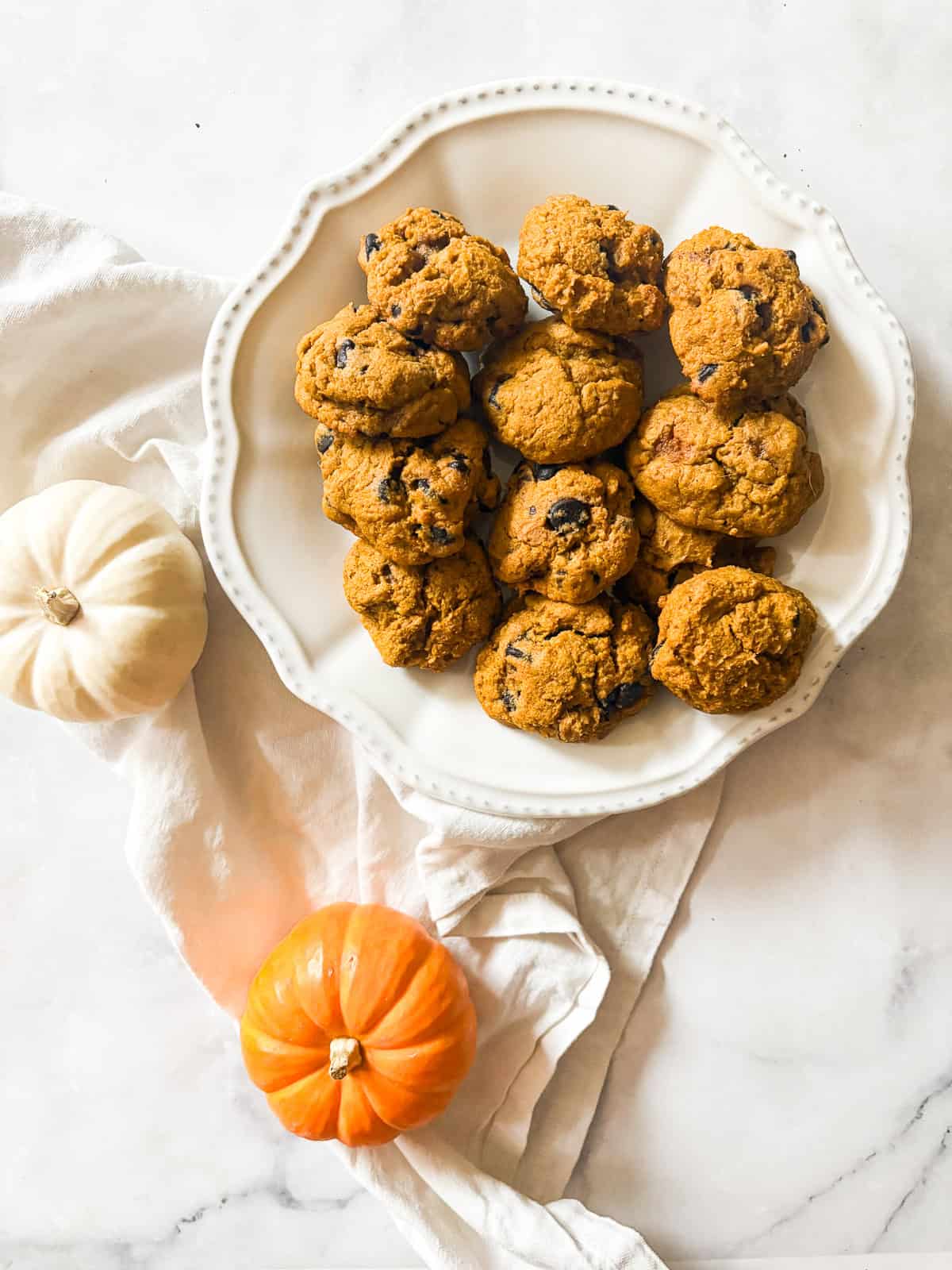 These soft-baked gluten free pumpkin chocolate chip cookies are packed with pumpkin puree, warm spices, and semisweet chocolate. Mini pumpkins and a plate of gluten free pumpkin chocolate chip cookies.