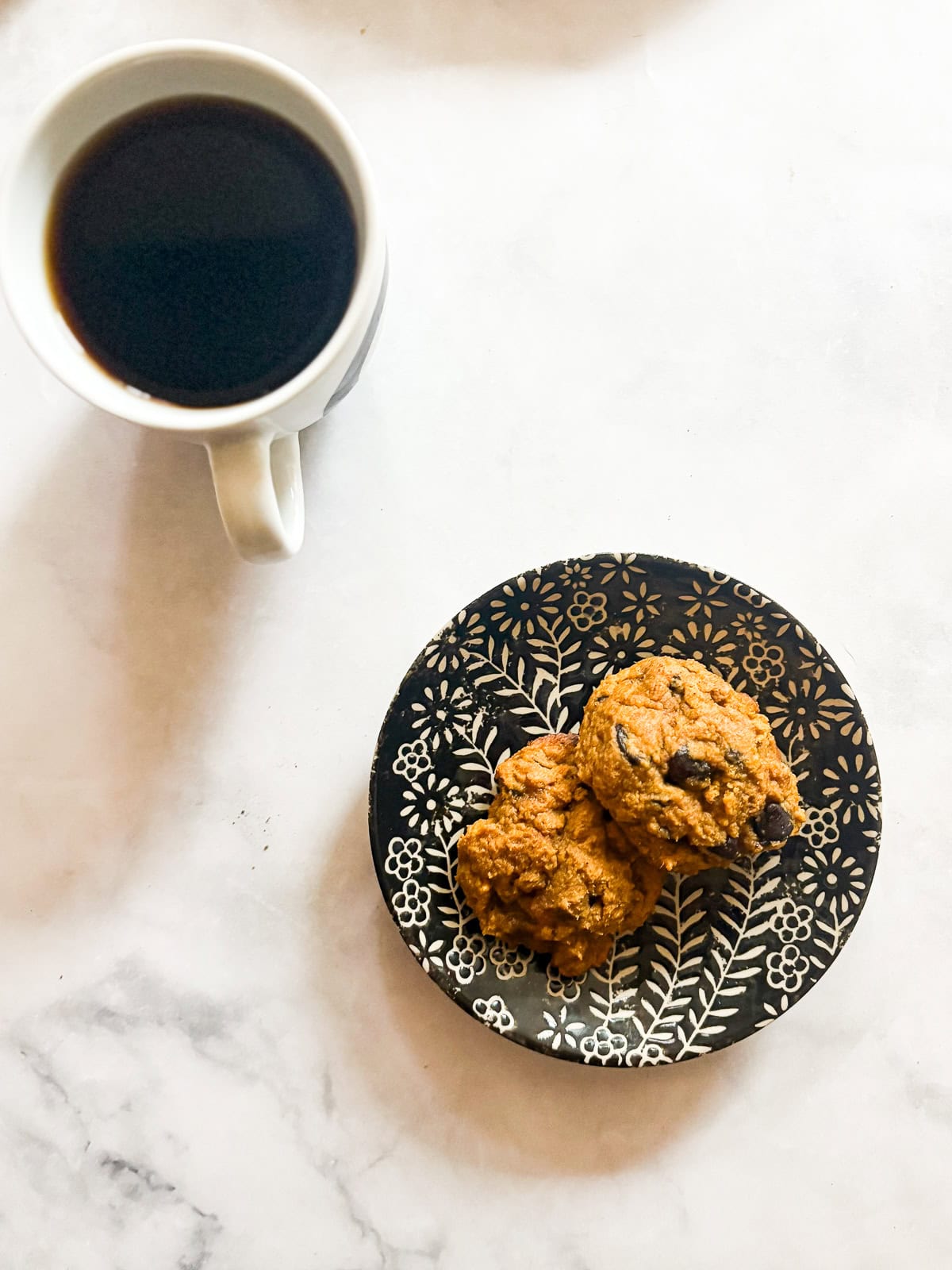 These soft-baked gluten free pumpkin chocolate chip cookies are packed with pumpkin puree, warm spices, and semisweet chocolate. A cup of coffee and two pumpkin chocolate chip cookies on a plate.