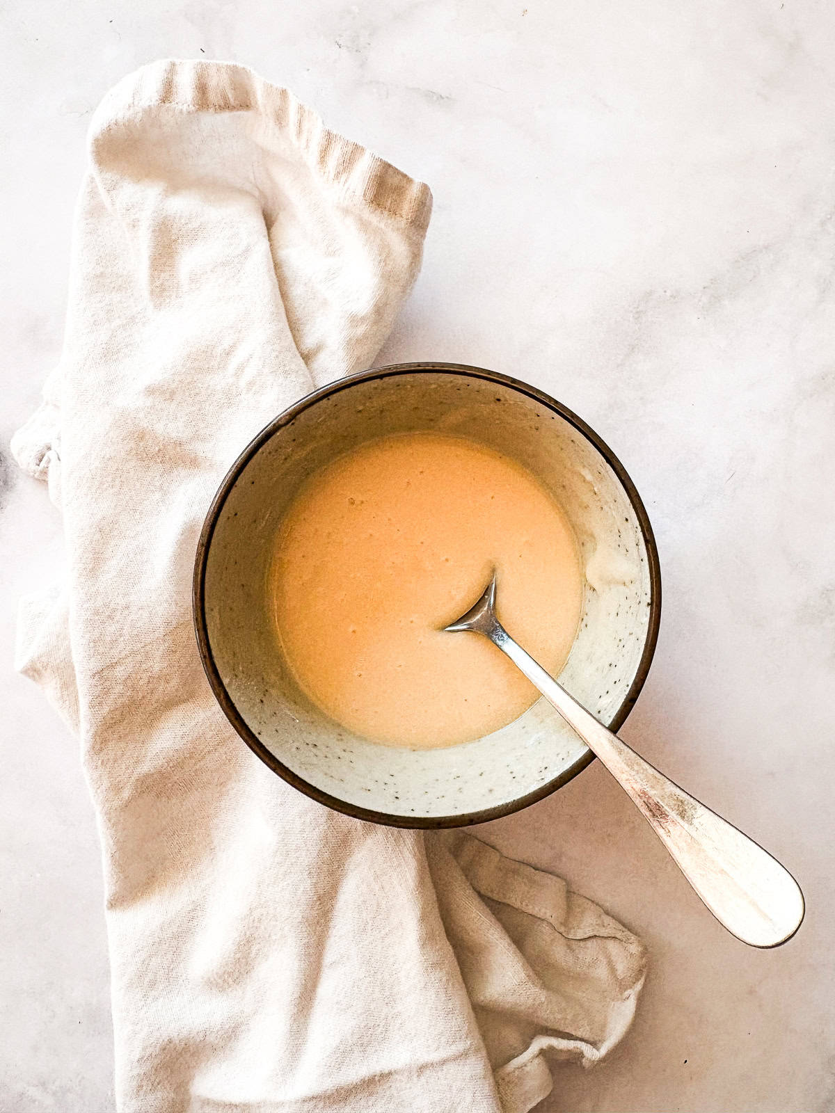 This smooth and luxurious maple glaze recipe needs just three simple ingredients to make and is wonderful on scones, pumpkin bread, and more. A bowl of maple glaze on a white napkin with a spoon.
