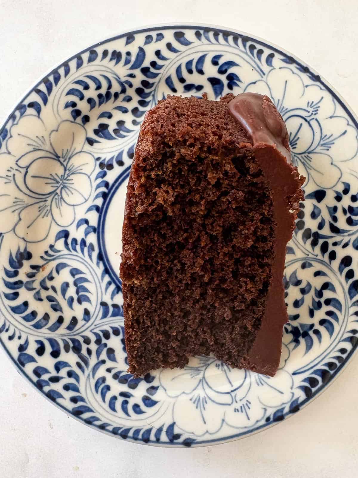 A tender, deeply dark gluten free chocolate olive oil cake capped with a layer of homemade chocolate icing. A slice of gluten chocolate olive oil cake on a blue and white plate.