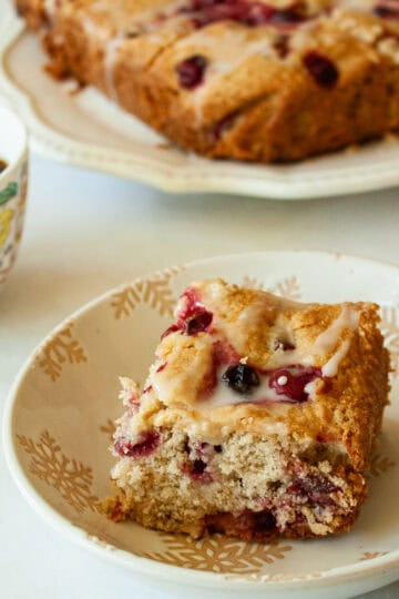 A cup of coffee next to a piece of gluten free cranberry cake with the cake in the background.