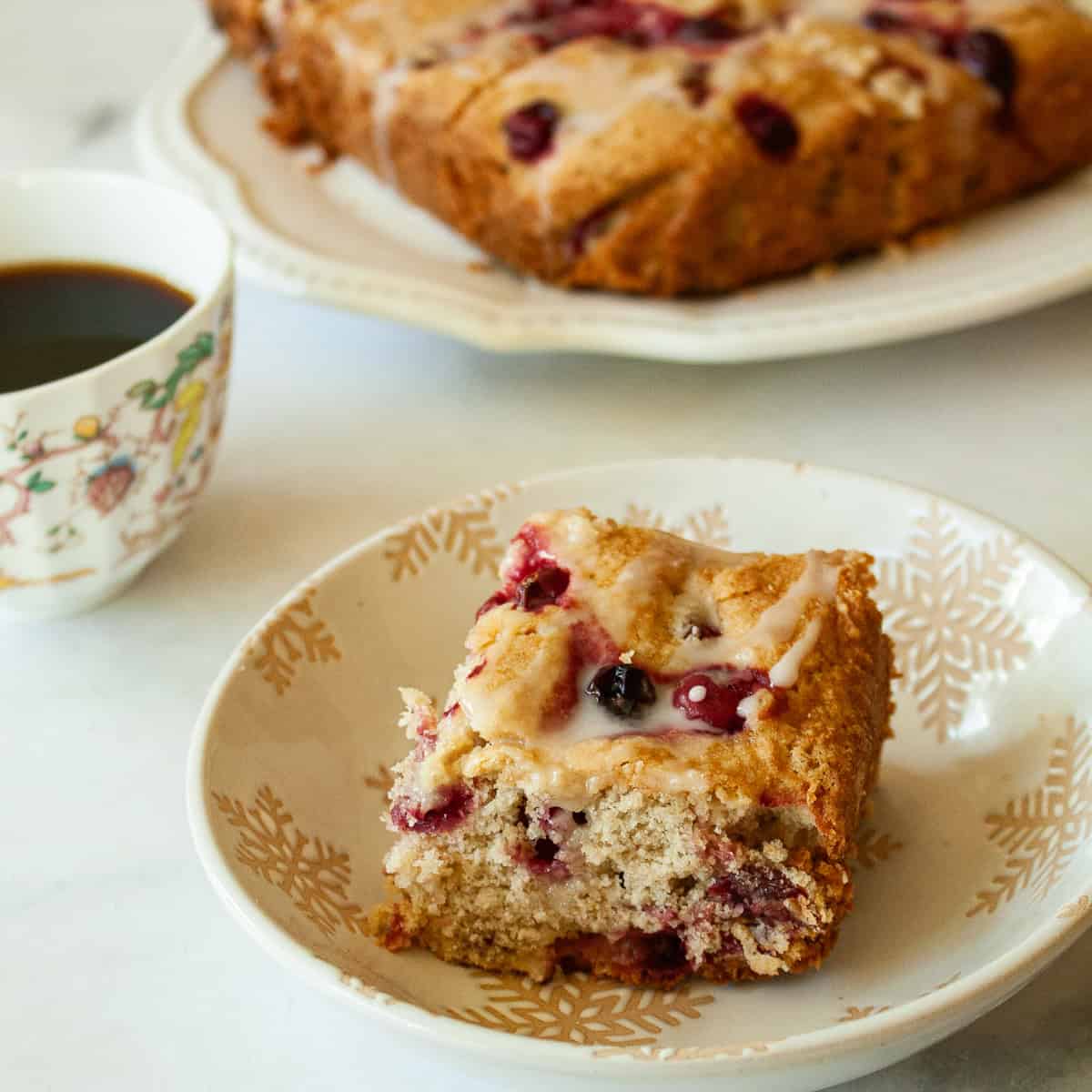This beautifully festive gluten free cranberry cake is packed with juicy, tart berries that are balanced by a sweet, buttery cake. A cup of coffee next to a piece of gluten free cranberry cake with the cake in the background.