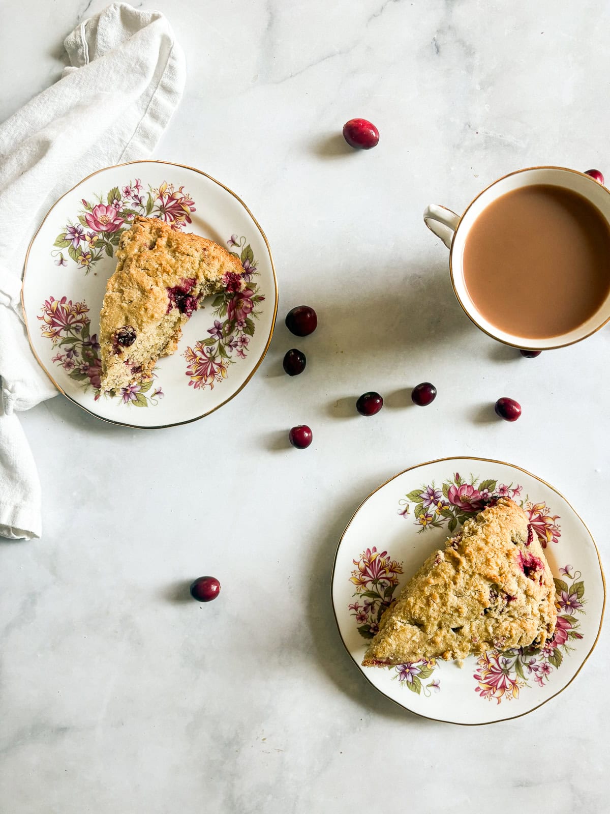 Tender and lightly sweet, these gluten-free cranberry scones are infused with orange zest and juicy cranberries. Two gluten free cranberry scones on plates, with cranberries, and a cup of tea.