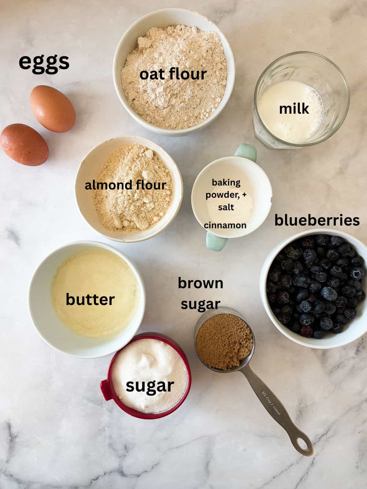 These cakey gluten-free bakery-style blueberry muffins are bursting with berries and topped with a cinnamon-spiked brown sugar crumble. The ingredients for gluten-free bakery-style blueberry muffins are text-labeled.