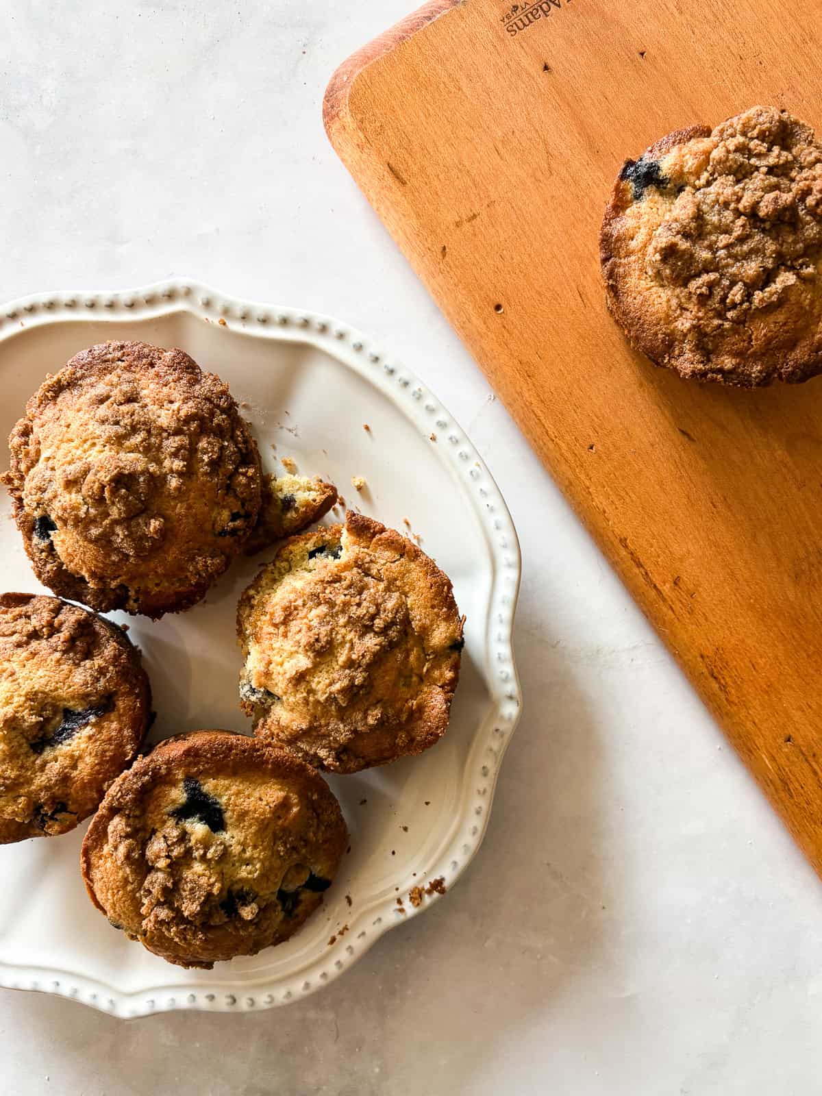 These cakey gluten-free bakery-style blueberry muffins are bursting with berries and topped with a cinnamon-spiked brown sugar crumble. A plate of bakery-style blueberry muffins next to a wooden board.