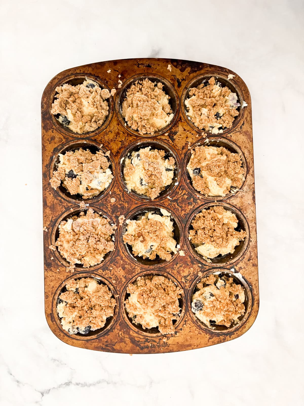 These cakey gluten-free bakery-style blueberry muffins are bursting with berries and topped with a cinnamon-spiked brown sugar crumble. Batter for gluten-free bakery-style blueberry muffins in a muffin tin.