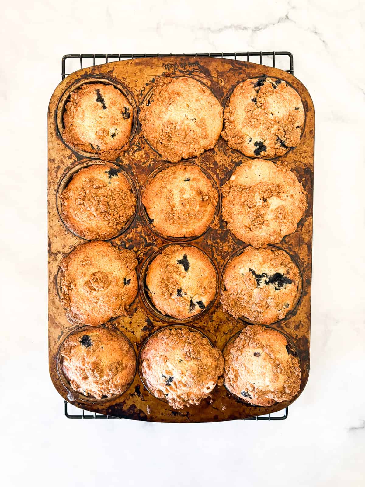 These cakey gluten-free bakery-style blueberry muffins are bursting with berries and topped with a cinnamon-spiked brown sugar crumble. Gluten free bakery-style blueberry muffins cool in the muffin pan.
