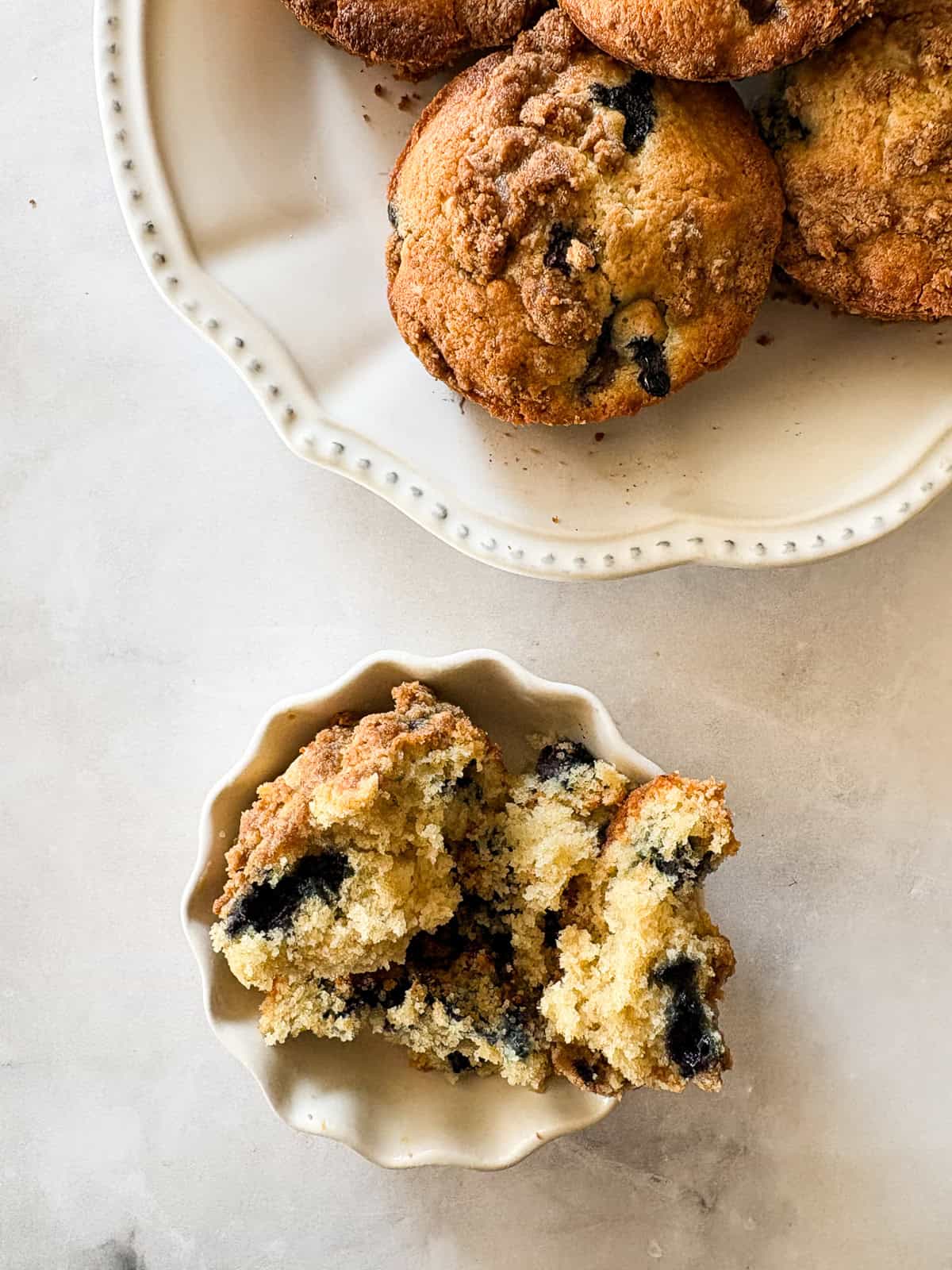 These cakey gluten-free bakery-style blueberry muffins are bursting with berries and topped with a cinnamon-spiked brown sugar crumble. A gluten-free bakery-style blueberry muffin split in half on a plate next to a plate of muffins.