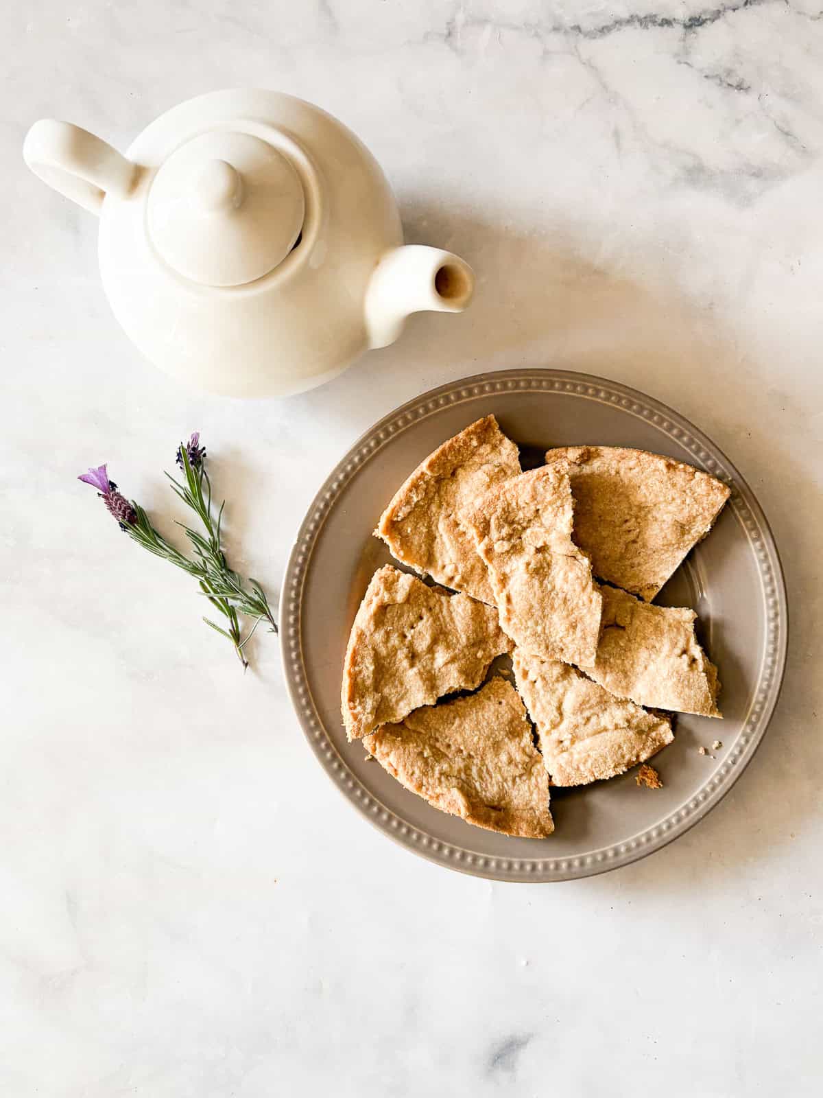 Melt-in-your-mouth gluten free lavender shortbread cookies are made with plenty of butter and infused with a bit of fresh or dried lavender. A white tea pot next to a plate of gluten free lavender shortbread cookies and a few sprigs of lavender.