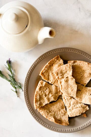 A white tea pot next to a plate of gluten free lavender shortbread cookies and a few sprigs of lavender.