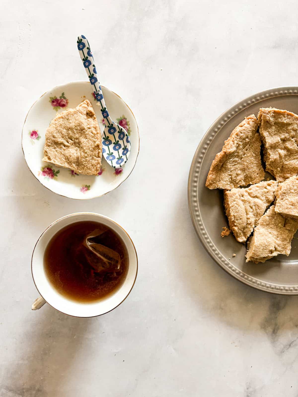 A spoon rests next to a gluten free lavender shortbread cookie, with a cup of tea, and a plate of cookies.