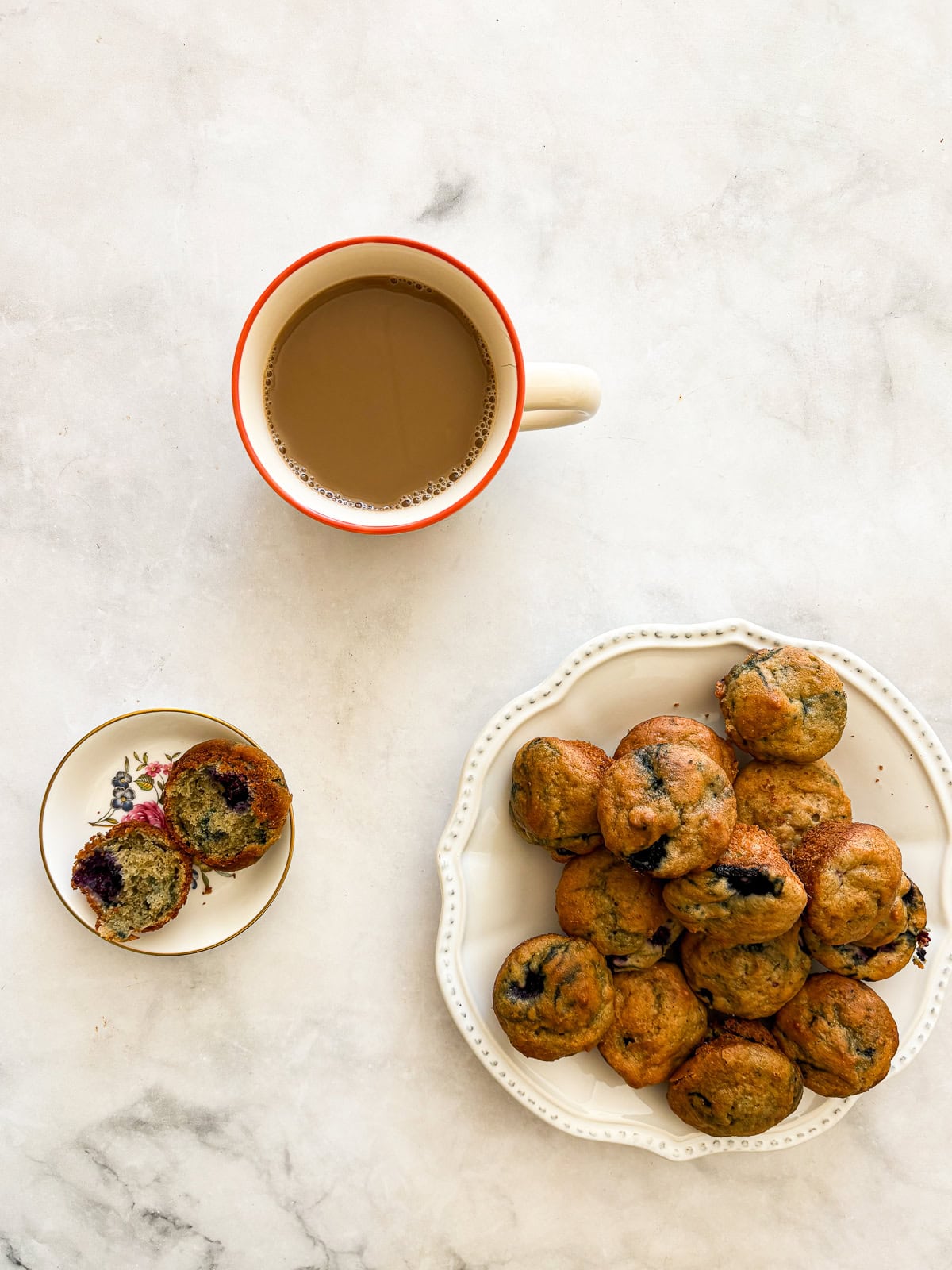 Two-bite, gluten-free mini blueberry muffins are made with whole-grain oat flour, Greek yogurt, and plenty of juicy berries. Kids love them! A mini blueberry muffin split in half on a plate next to a plate of muffins and a cup of coffee.
