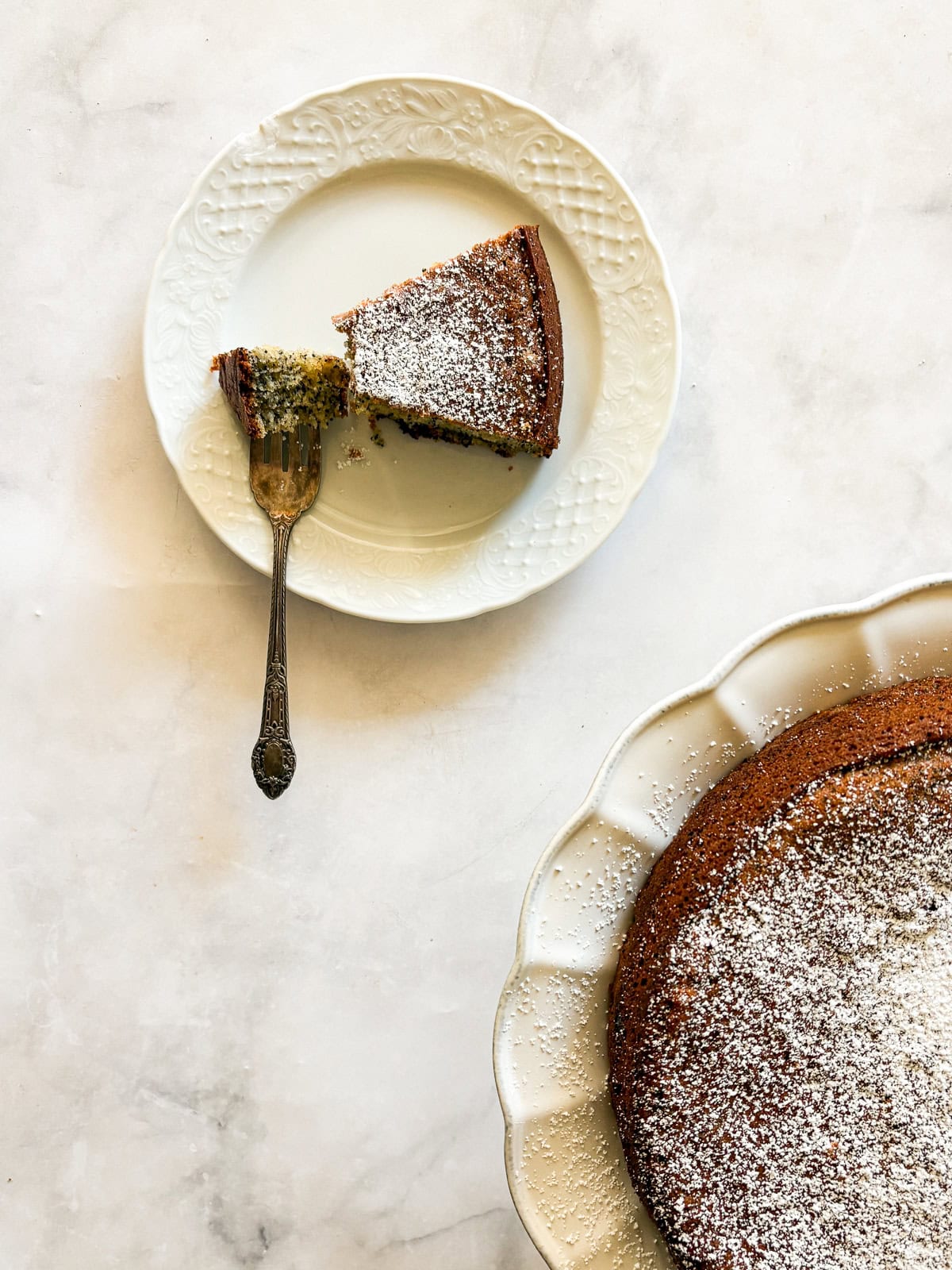 his gluten-free poppyseed cake is tender-crumbed, generously speckled with poppyseeds, and made with two simple flours. A fork cuts into a piece of gluten free poppyseed cake on a plate next to the cake.
