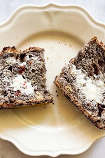 A slice of gluten free no knead cranberry walnut bread cut in half and spread with butter on a plate.