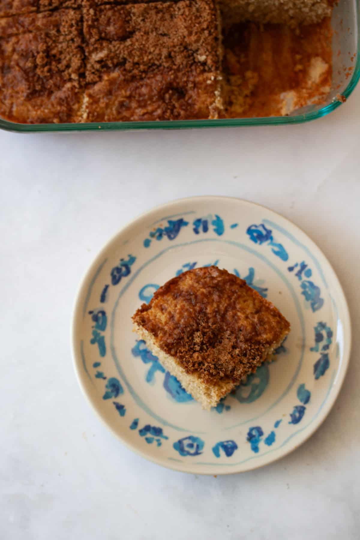 This super easy gluten free cinnamon and sugar cake is perfect for a last-minute dessert or quick breakfast coffee cake option! A slice of gluten free cinnamon and sugar cake on a plate with the pan of cake behind it.