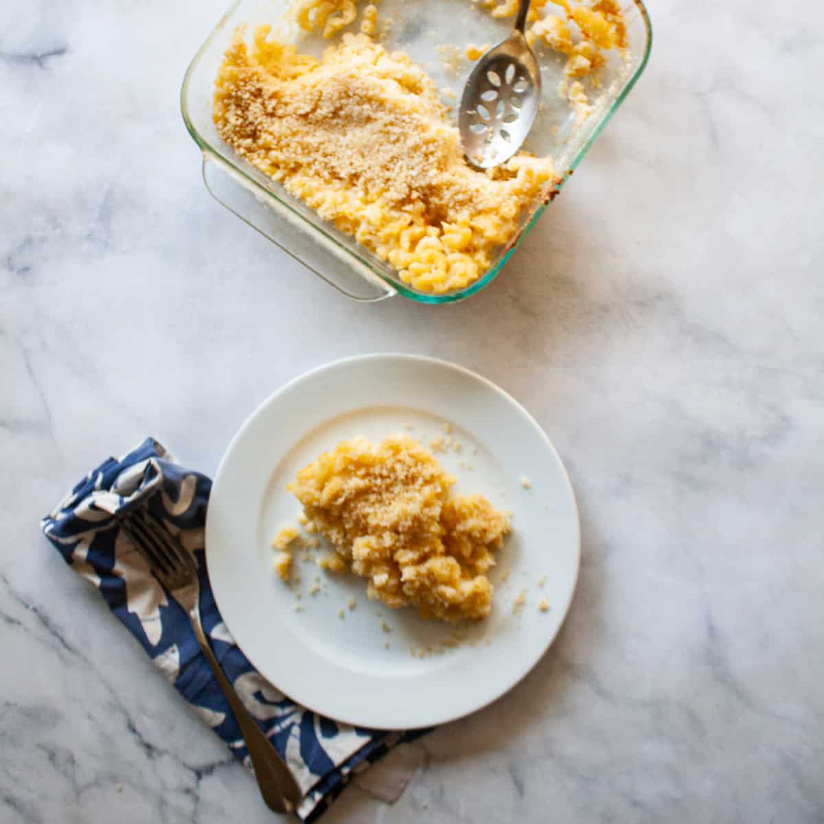 A serving spoon in a pan of kids mac and cheese next to a plate of mac and cheese.