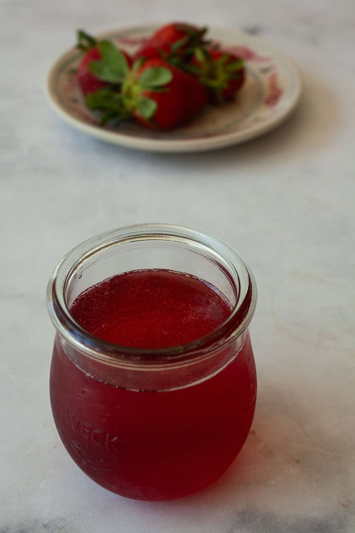 Three-ingredient strawberry simple syrup is so pretty when stirred into drinks or draped over cakes! A summer must-make. A plate of strawberries behind a jar of strawberry simple syrup.