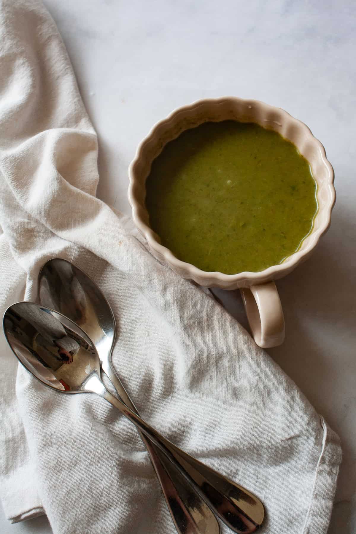 Creamy, vegan asparagus and spinach soup is made with minimal ingredients to create a velvety, nourishing soup that's always welcome. A cup of asparagus and spinach soup next to a white napkin with spoons on it.