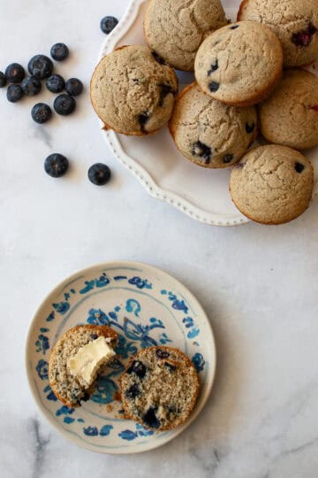 Blueberries, a plate of whole grain gluten free berry muffins, and a muffin with butter on a plate.