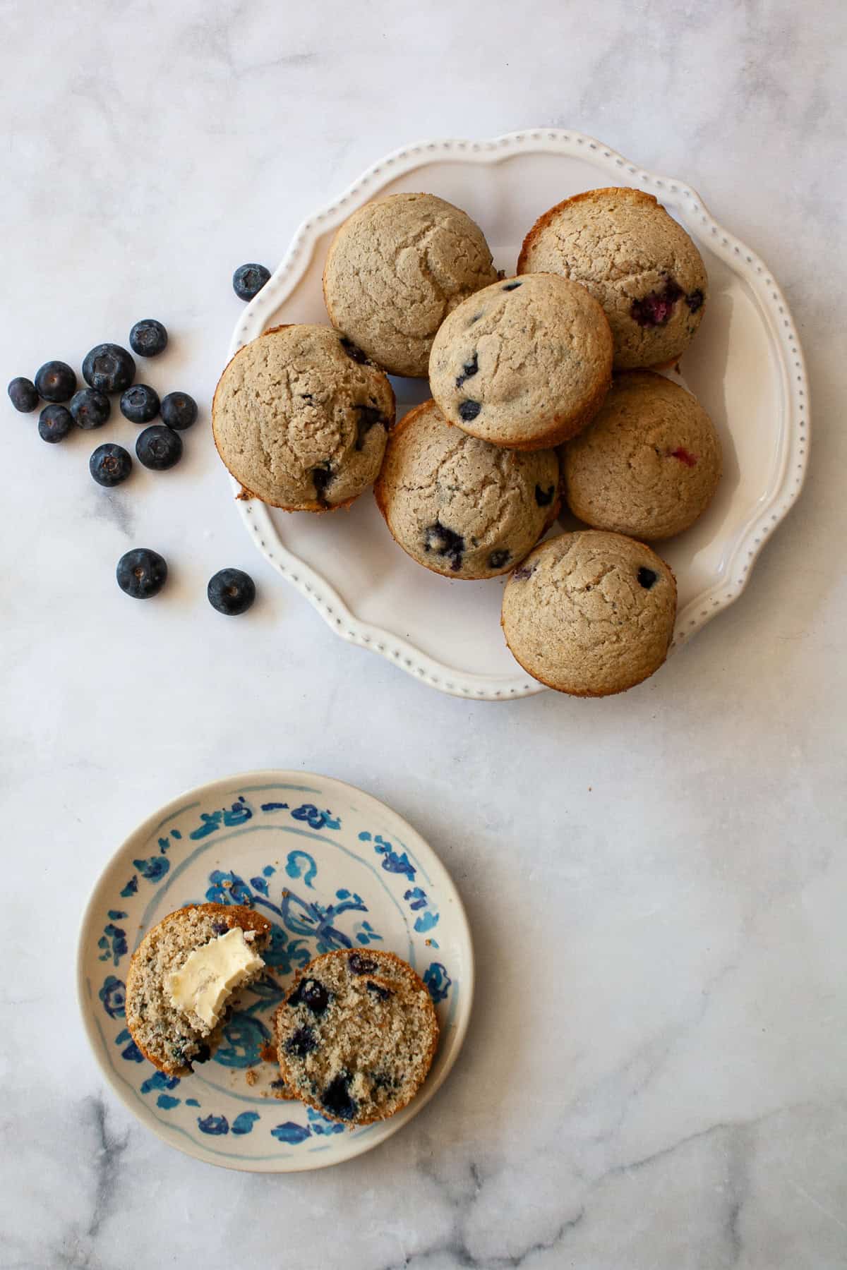 Blueberries, a plate of whole grain gluten free berry muffins, and a muffin with butter on a plate.