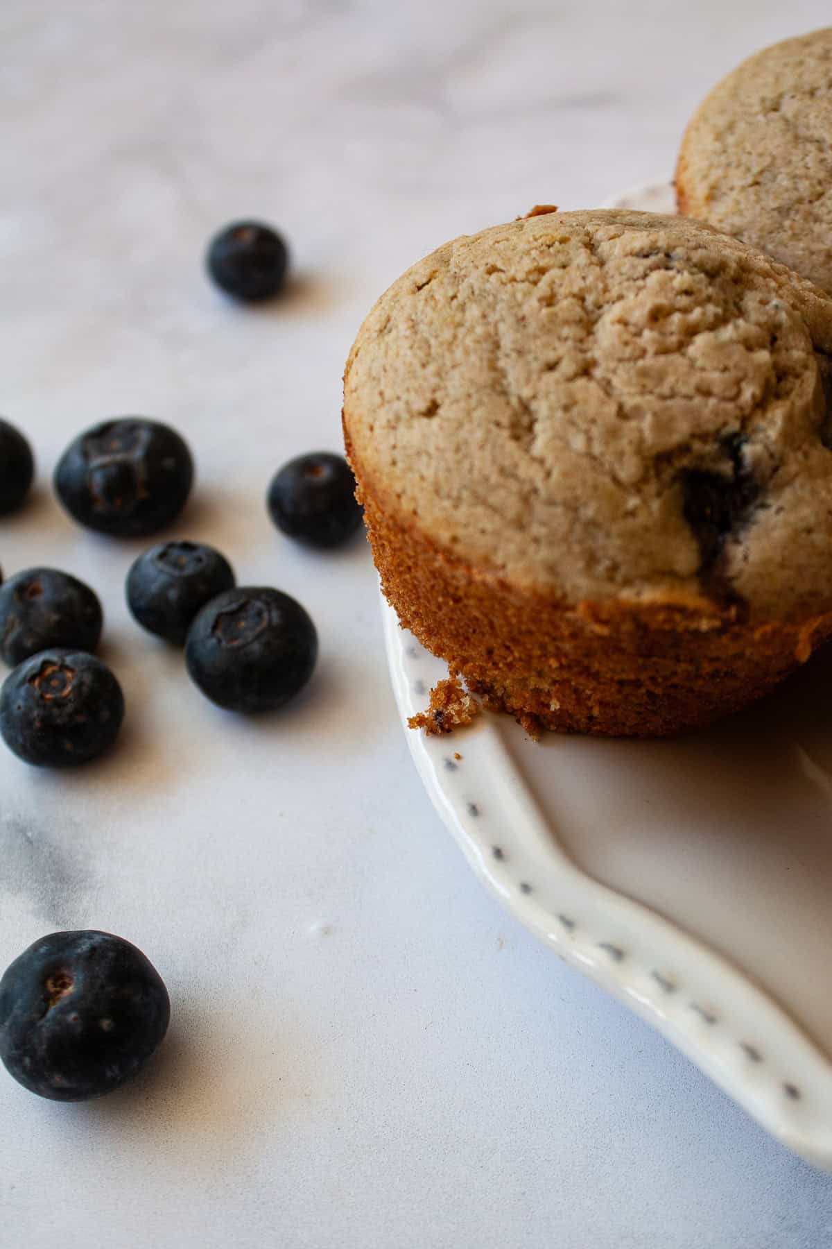These one-bowl gluten-free berry muffins are made with your choice of whole grain flour and are sweetened just with maple syrup! Blueberries scattered near a gluten free berry muffin.