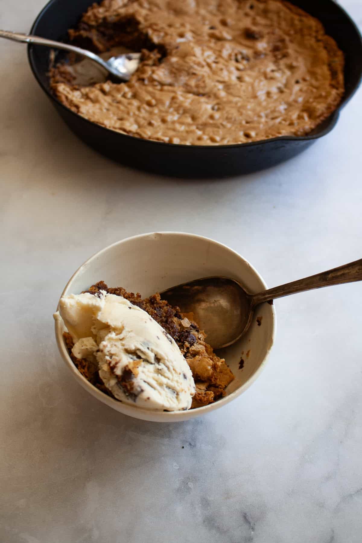 This sweet and chewy gluten free skillet cookie that's soooo good served warm and topped with ice cream. Made in one bowl! A portion of gluten free skillet cookie in a bowl topped with ice cream with the skillet behind it.