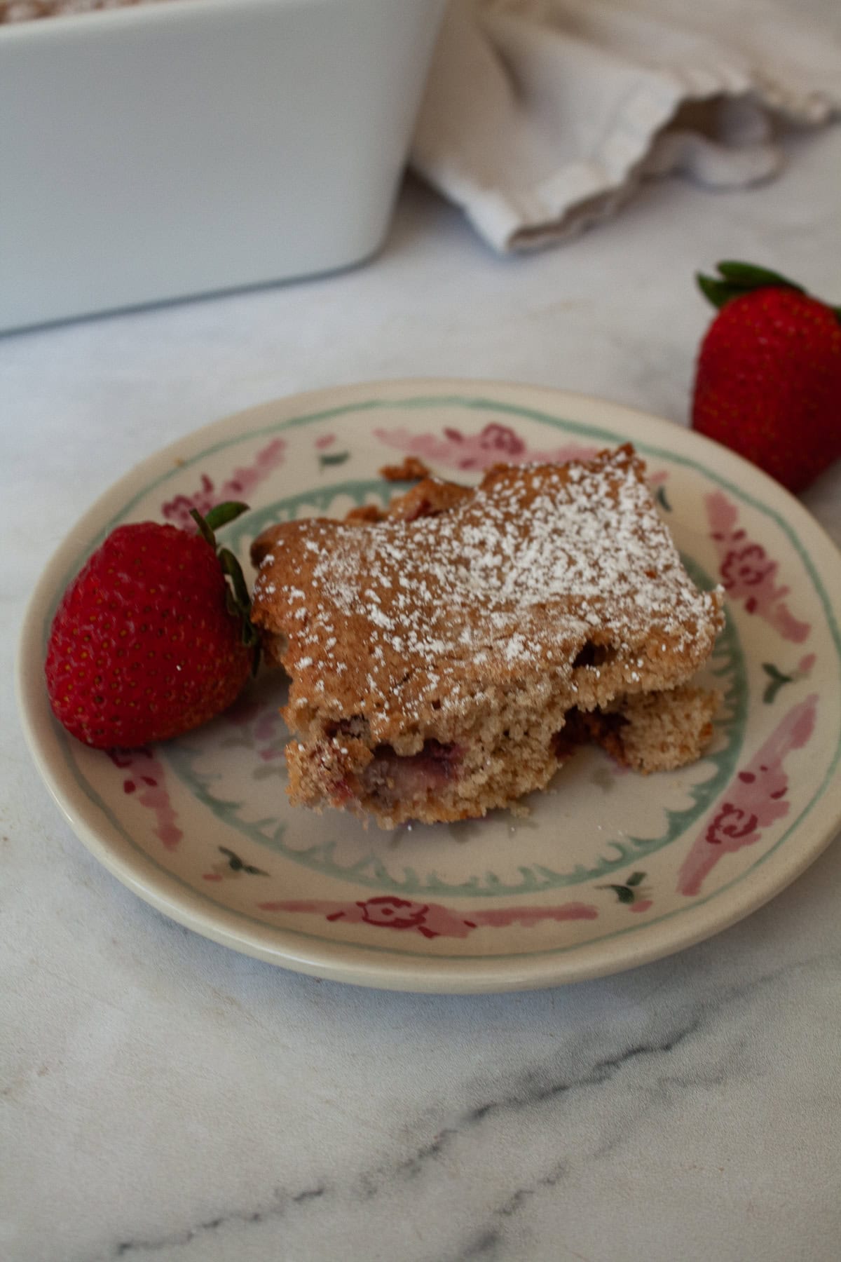 A strawberry next to a slice of gluten free strawberry rhubarb cake on a plate.
