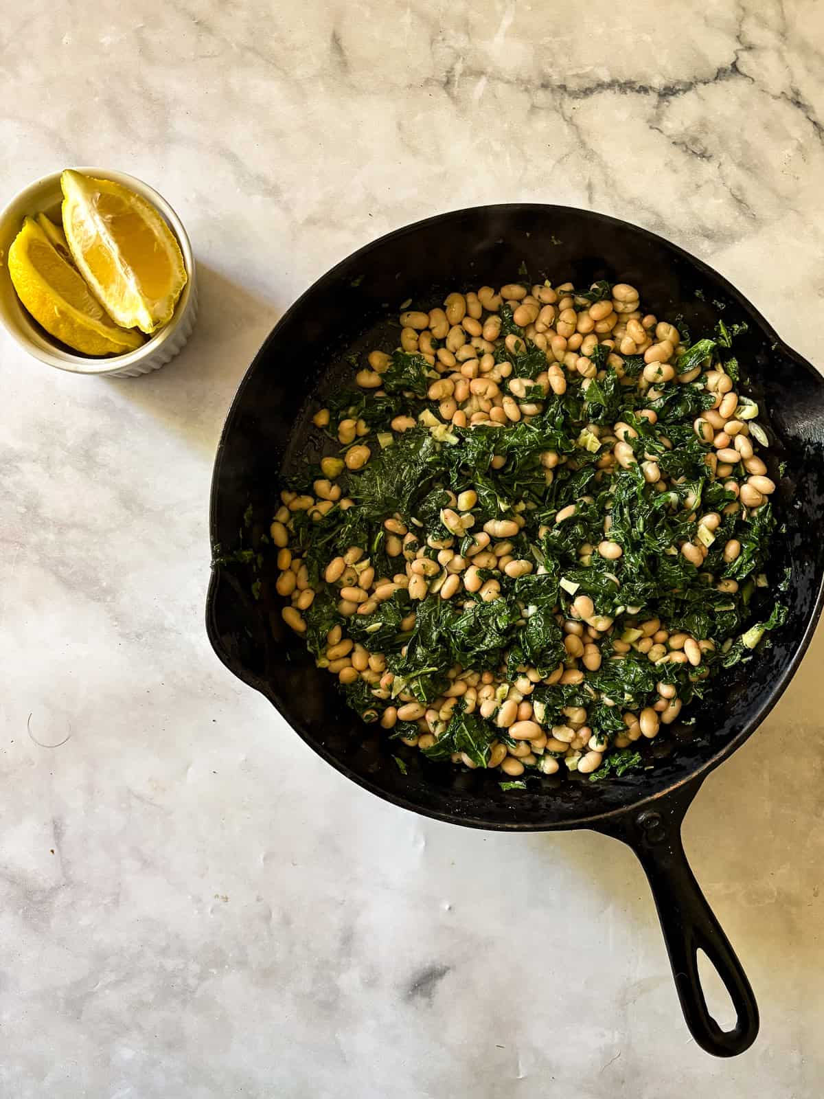 A skillet of white beans and kale with lemon wedges next to it.