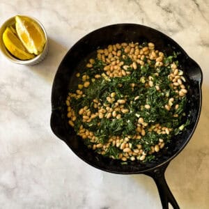 A skillet of white beans and kale with lemon wedges next to it.