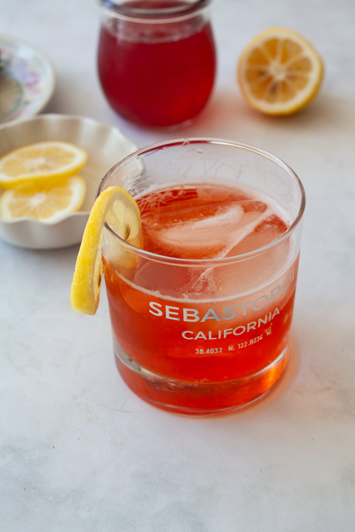Enjoy this strawberry gin and tonic all summer long! A fruit twist on a classic, sweet strawberry syrup elevates this familiar cocktail. Lemons, strawberry syrup in the background and a glass of strawberry gin and tonic in the front.