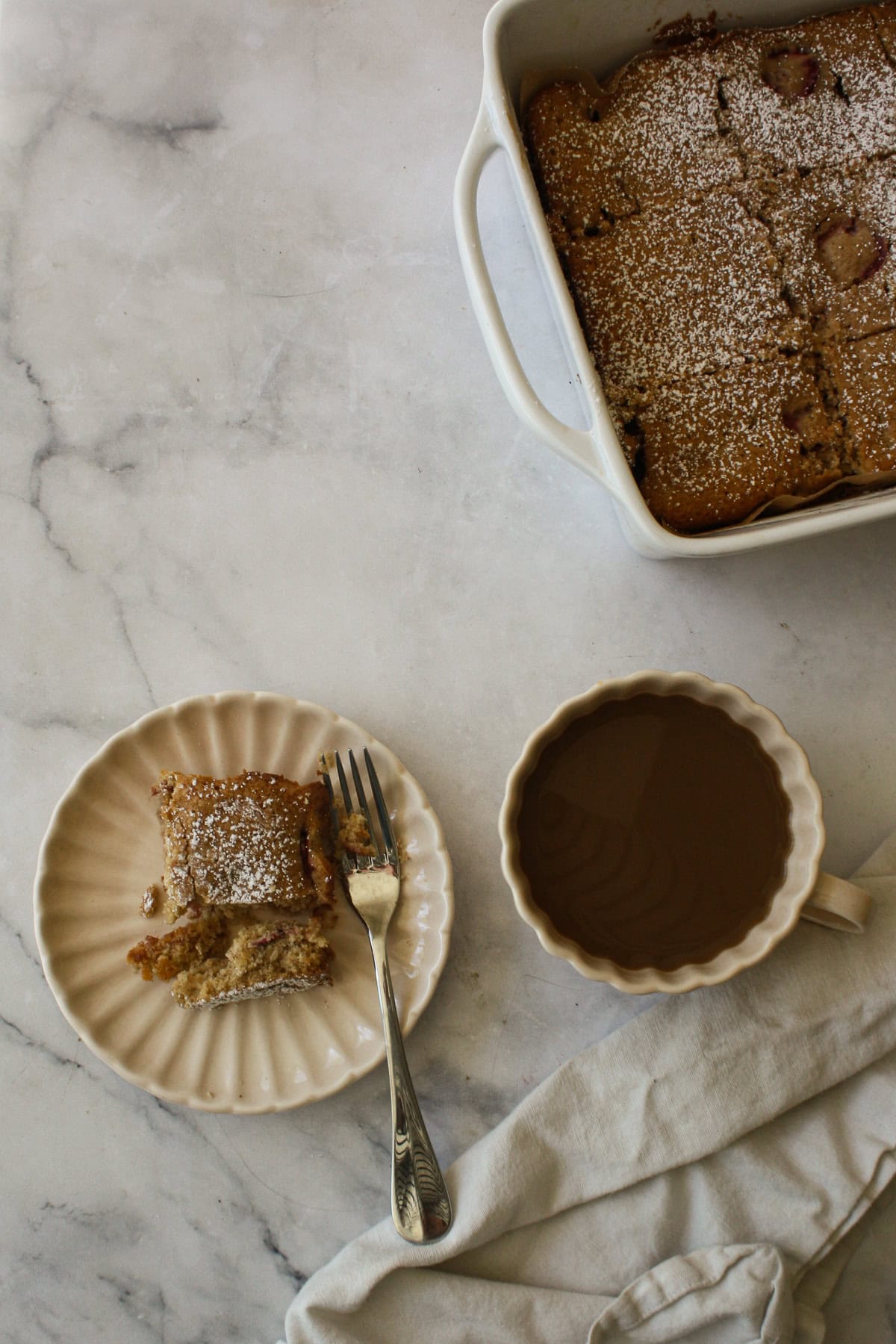 A pan of strawberry rhubarb cake, a coffee, and a slice on a plate.