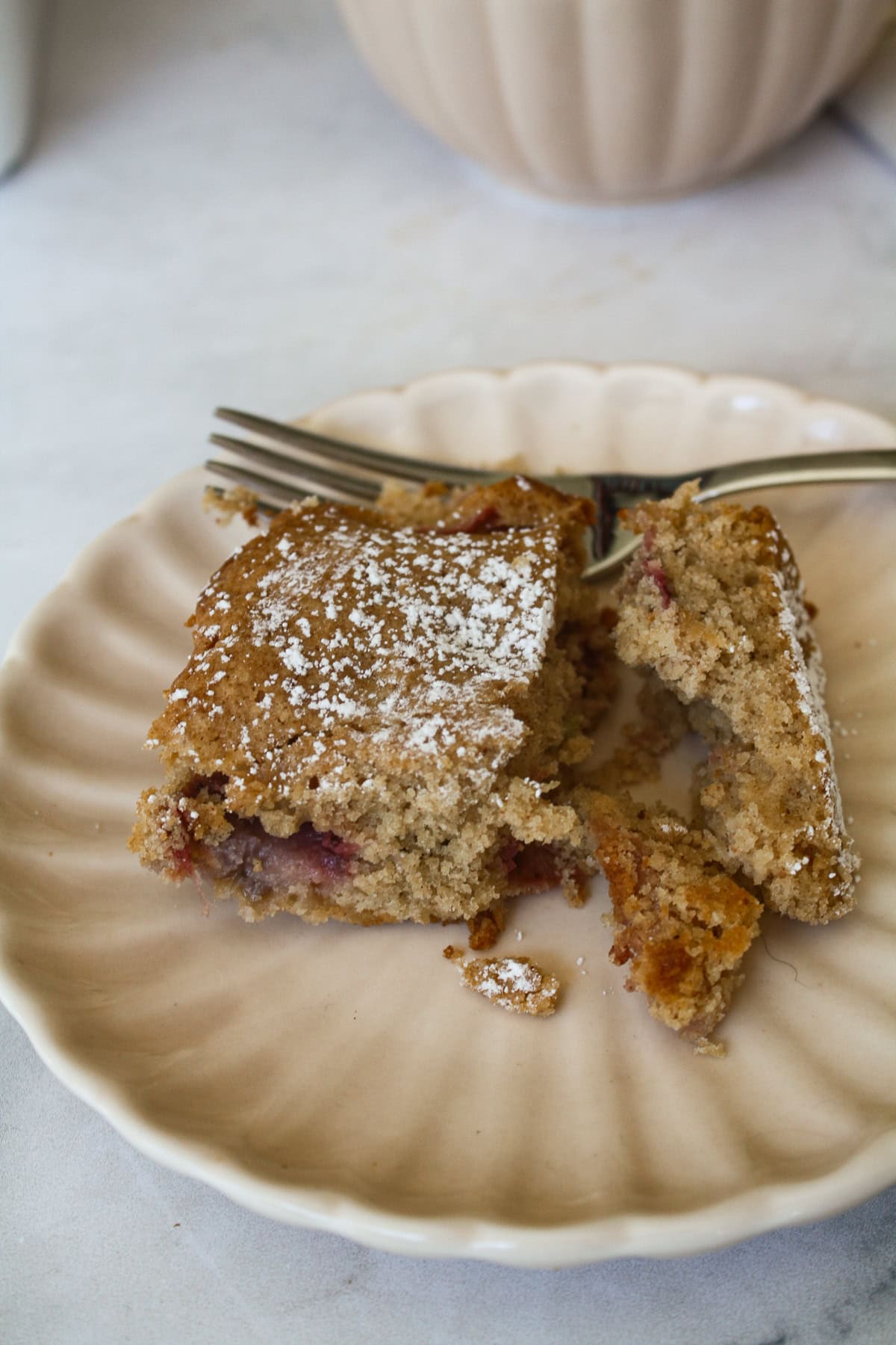 A tender strawberry rhubarb cake made with whole wheat flour, fresh fruit, and a buttery crumb. Rustic, easy, and perfect for summer baking. A cup of coffee behind a plate with strawberry rhubarb cake.