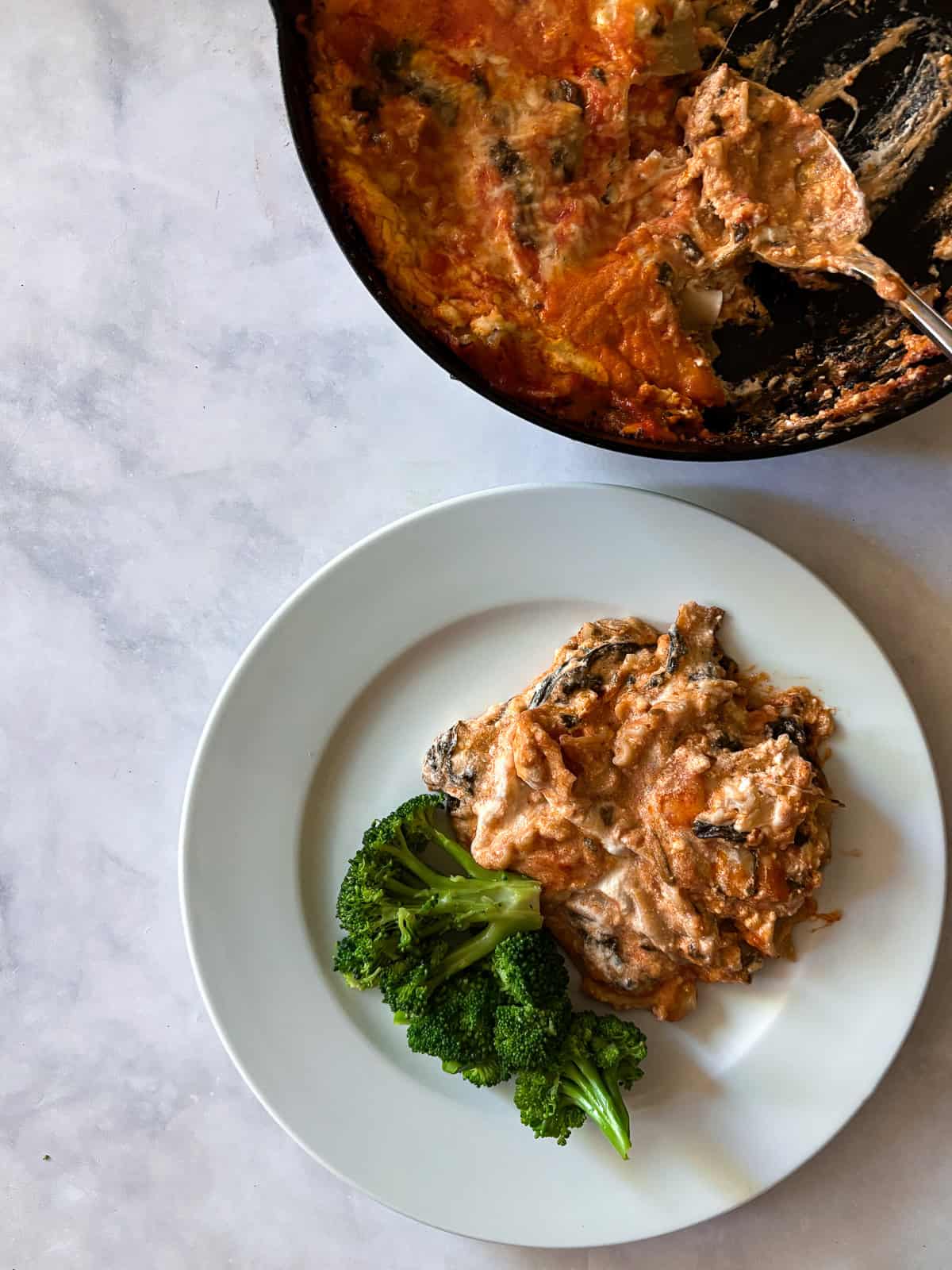 Broccoli and a serving of gluten free skillet lasagna on a plate with the pan of lasagna next to it.