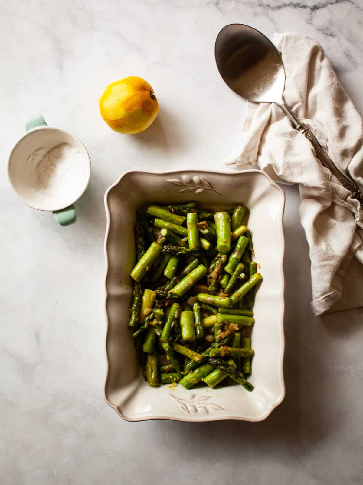 A serving dish with asparagus and lemon plus a lemon, salt, and a serving spoon.