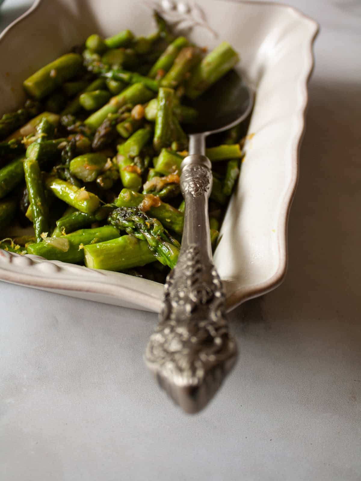 Cook to taste. I love that sweet spot between cooked through and still firm. You can cook the asparagus a bit more if you prefer softer asparagus and conversely, can cook it a little less for firmer asparagus. Herbs. Stir in some chopped fresh or dried herbs to the pan, such as parsley, basil, or oregano. Add more garlic. This recipe takes well to adjustments. Add more garlic if you like a lot of garlic. You can also use less. Serve cold. While I tend to serve this sautee with a hot meal, it's also lovely cold! You could mix it with quinoa and chickpeas to make a warm-weather side dish, or add leftover asparagus to a salad. A serving spoon rests in a dish of asparagus and lemon.
