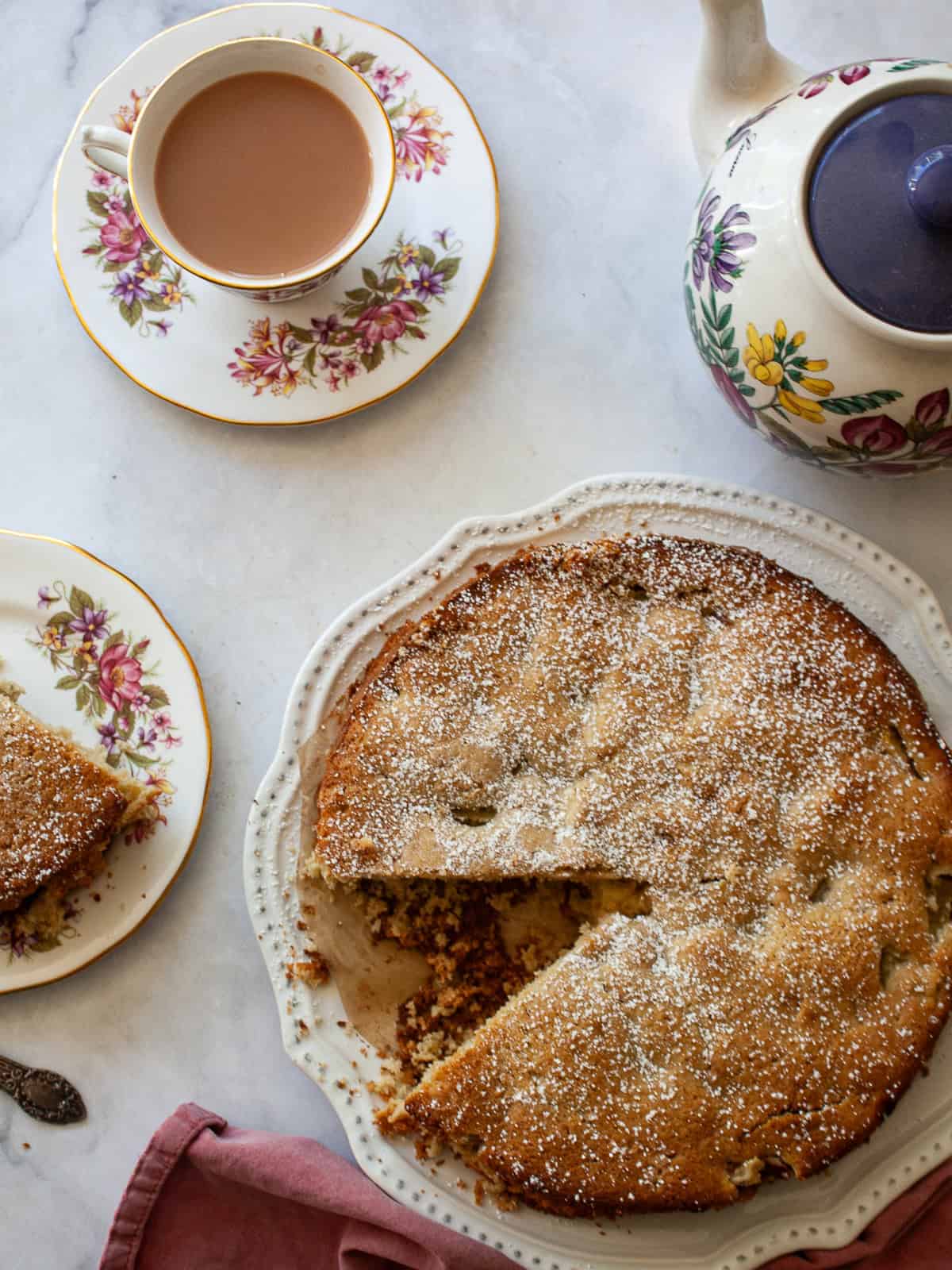 This pretty cake marries crisp, sweet apples with tart rhubarb to make a seasonal gluten free apple and rhubarb cake! A cup of tea, a tea pot, and a gluten free apple and rhubarb cake.