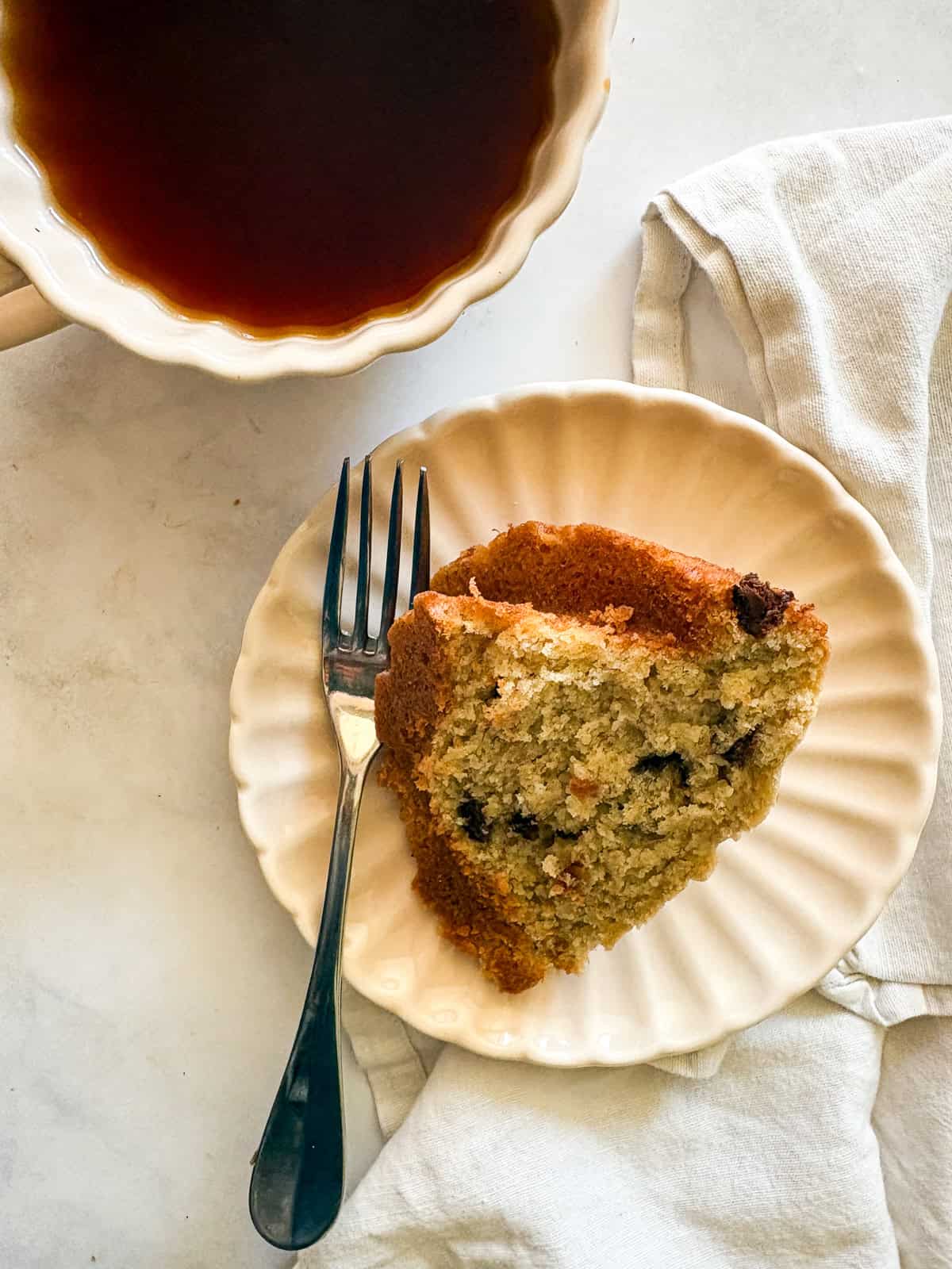 This rich, gluten free banana bundt cake is made with plenty of butter and sugar, then swirled with bananas and chocolate chips. A slice of gluten free banana bundt cake on a plate with a fork next to a cup of coffee.