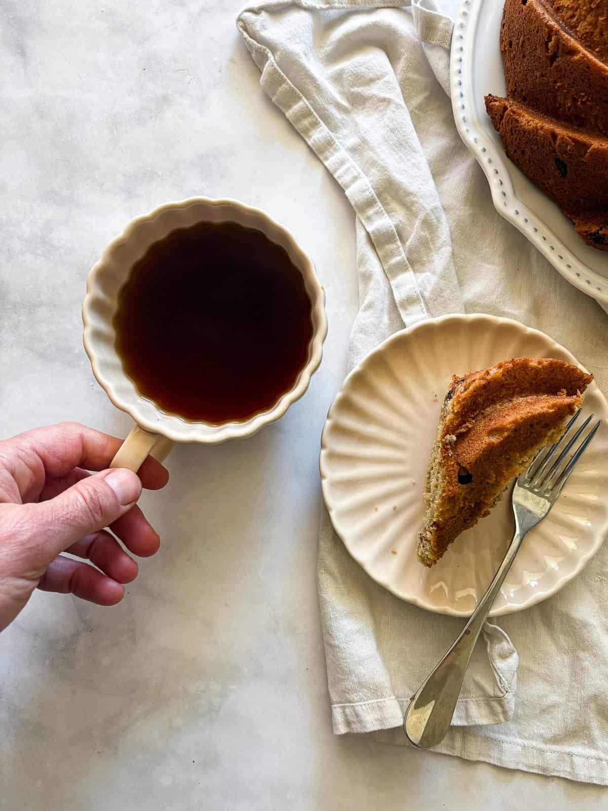 This rich, gluten free banana bundt cake is made with plenty of butter and sugar, then swirled with bananas and chocolate chips. A hand reached for a cup next to a banana bundt cake.