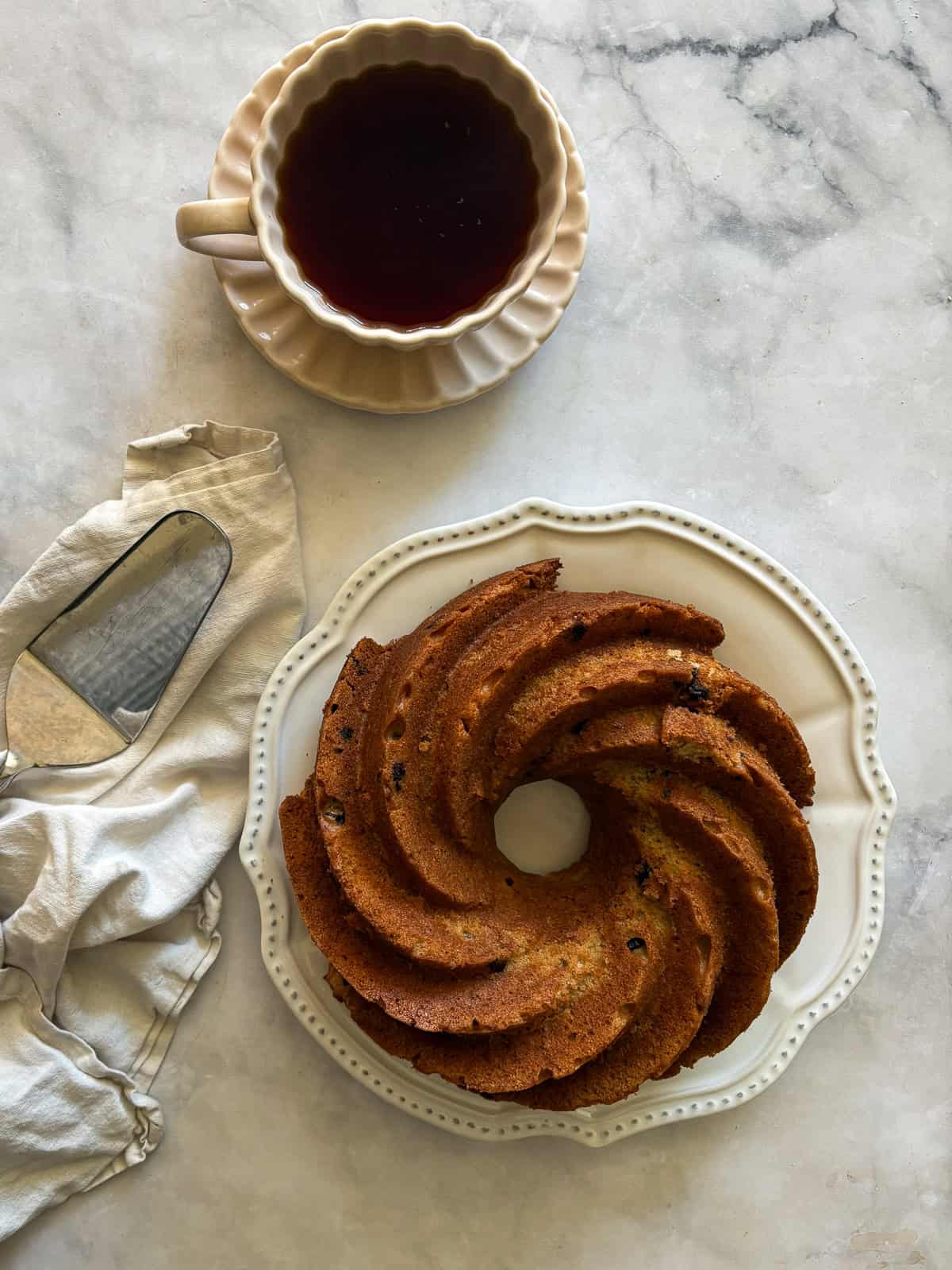 This rich, gluten free banana bundt cake is made with plenty of butter and sugar, then swirled with bananas and chocolate chips. A gluten free banana bundt cake on a plate next to a serving and a cup of coffee.