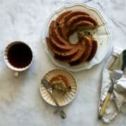 A slice of gluten free banana bundt cake on a plate with the cake and a cup of coffee.