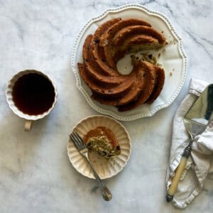 A slice of gluten free banana bundt cake on a plate with the cake and a cup of coffee.