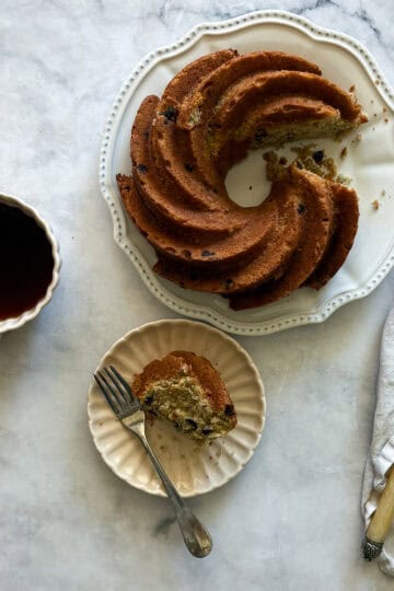 A slice of gluten free banana bundt cake on a plate with the cake and a cup of coffee.